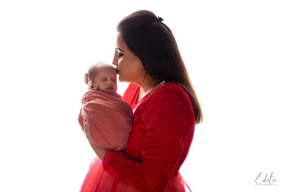 Mother in red outfit kissing newborn baby girl, backlit photo in Pune